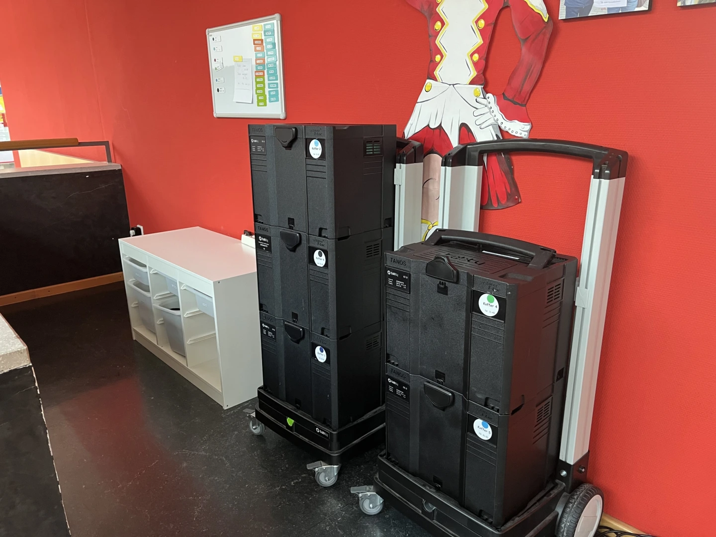 Zu sehen ist ein sogenannter Tabletkoffer. Dieser ist in einem Gang an die Wand geschoben und besteht aus zwei schwarzen Plastikkoffern, die aufeinander gestapelt auf einer Halternund mit Griff und Rollen stehen.