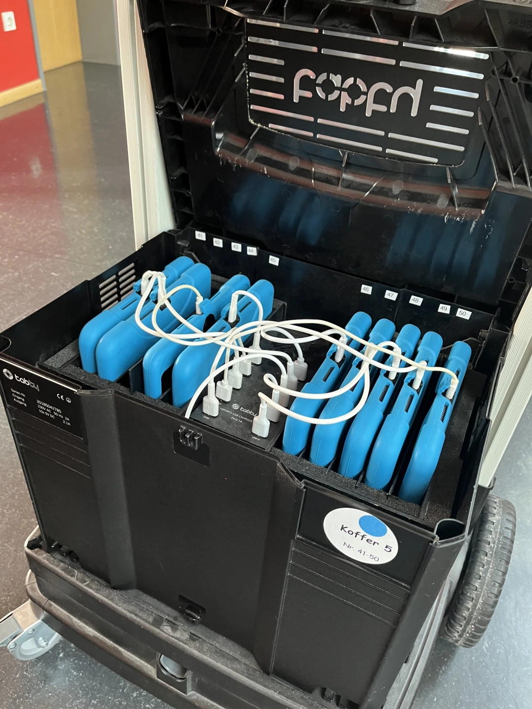 Ein Tabletkoffer ist geöffnet, man sieht acht iPads, die in entsprechende Halterungen nebeneinander geschoben sind und mit Kabeln mit einer Ladevorrichtung in der Mitte des Koffers verbunden sind.