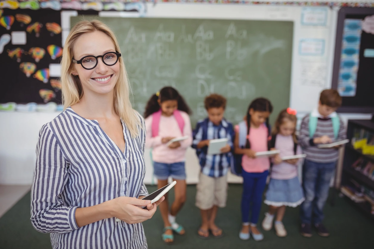 Lehrerin hält ein Tablet in der Hand, im Hintergrund stehen Schülerinnen und Schüler mit Tablets