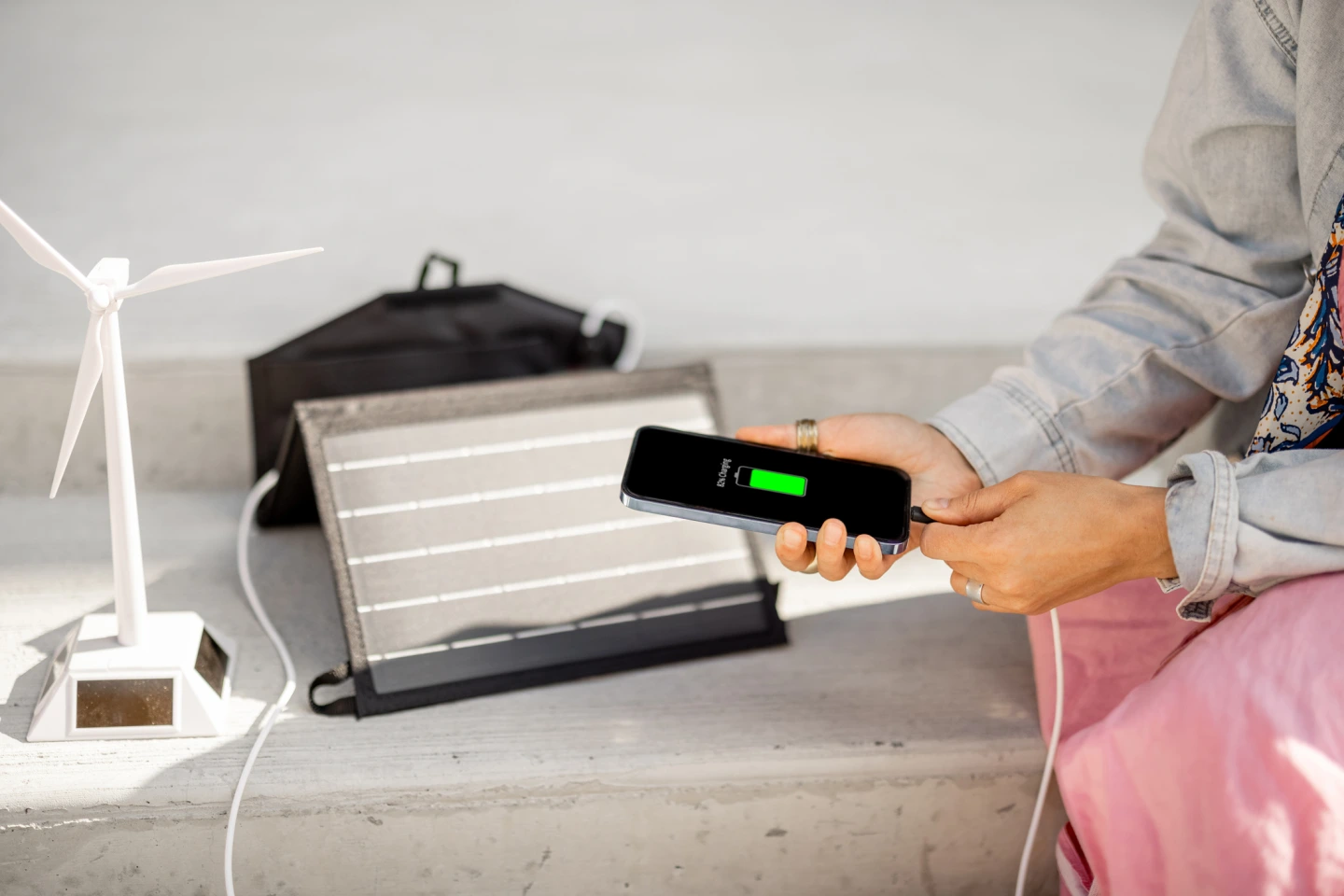 Eine Person steckt den Ladestecker eines Smartphones ein, das mit einem kleinen Windrad und einem tragbaren Solarpanel geladen wird.