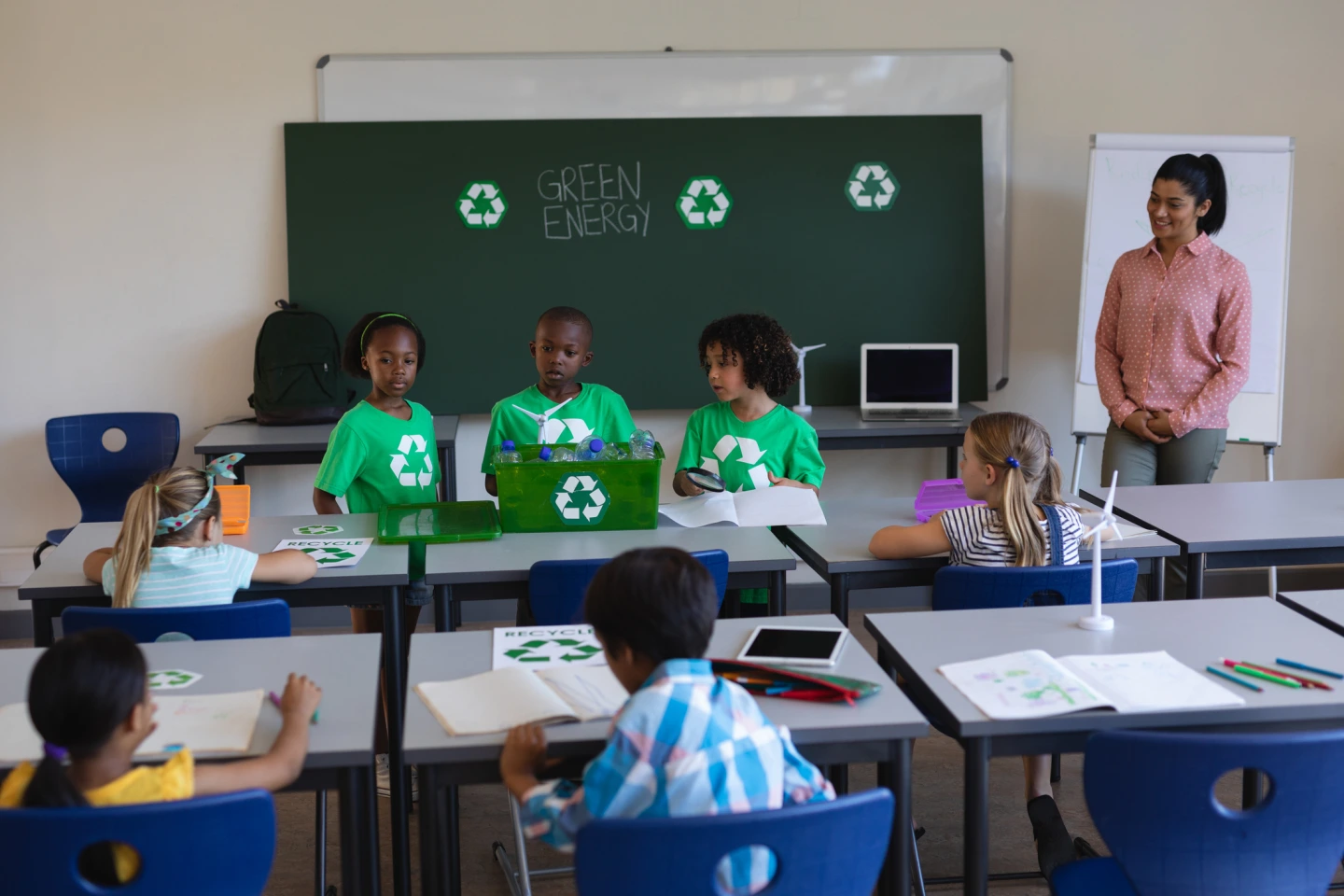 Drei Schülerinnen und Schüler in grünen T-Shirts mit aufgedrucktem weißen Recycling-Logo stehen in einem Klassenraum vor einer Tafel und halten einen Vortrag über Green Energie. Die Lehrerin und die Mitschüler hören aufmerksam zu.